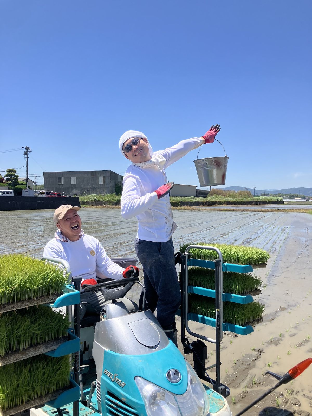 田植え機の上でバケツを掲げて笑う白石と、運転席で笑う仲間